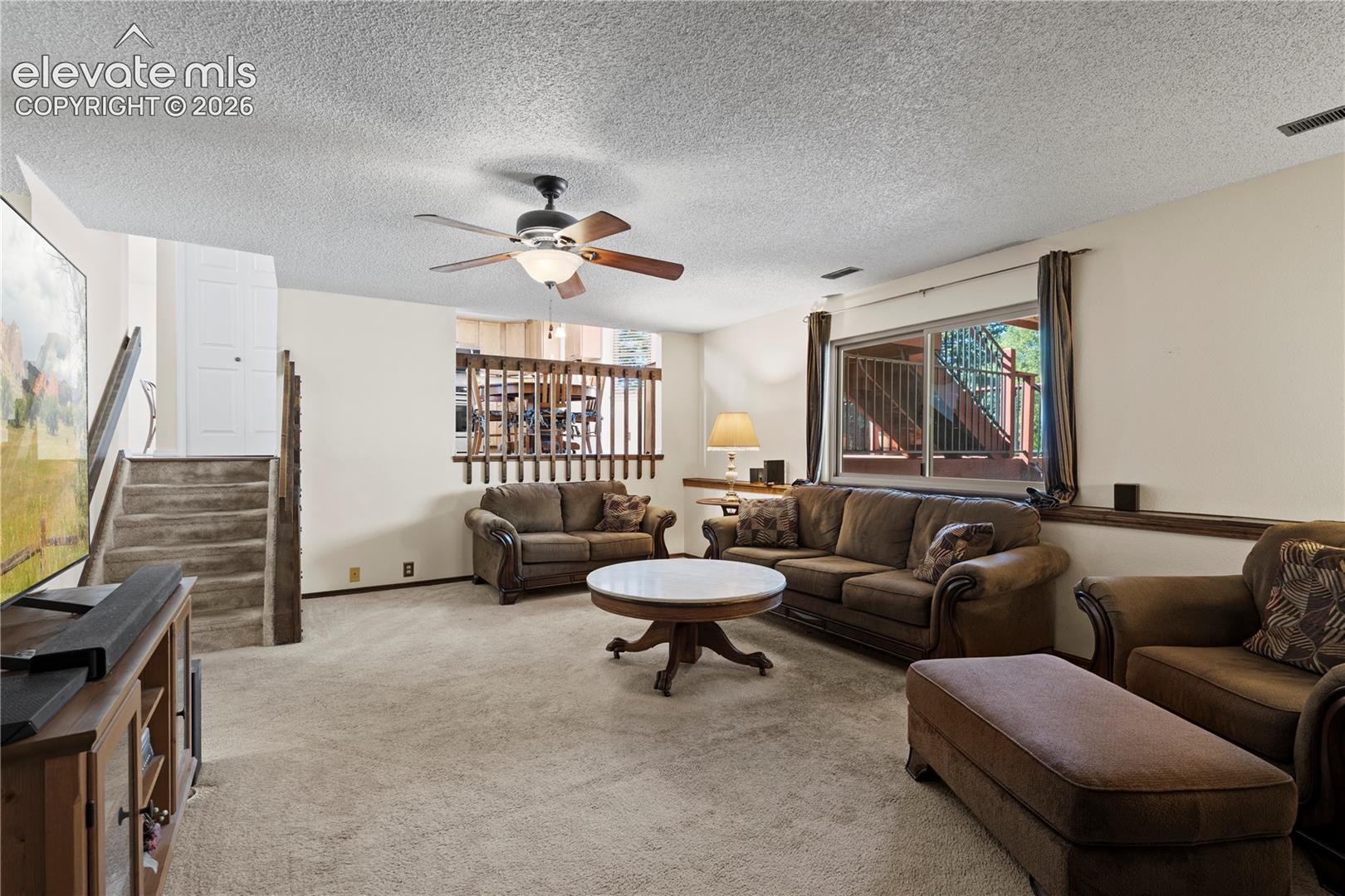 Carpeted living area featuring a ceiling fan, a prominent window, and a wood-finish staircase with railing