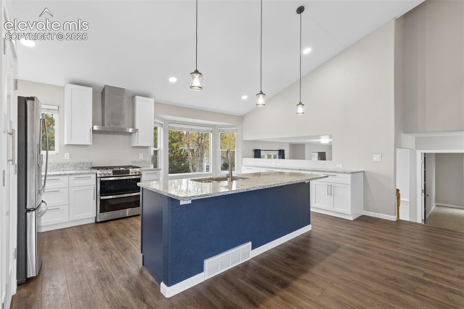 Kitchen featuring a central island with stone-finish countertops, stainless steel appliances, white shaker-style cabinetry, pendant lighting, and wood-finish flooring under a vaulted ceiling