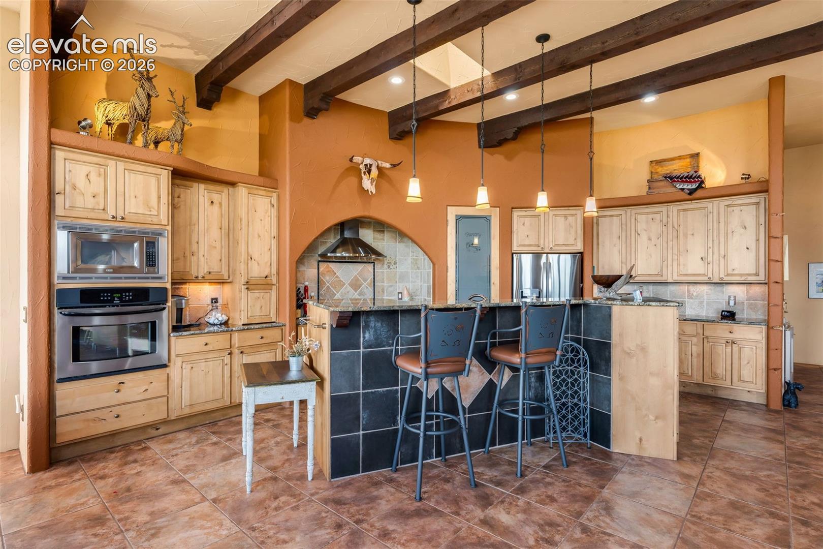 Kitchen featuring natural finish wood cabinetry, exposed ceiling beams, tile flooring, stone-tiled breakfast bar, and stainless steel built-in oven and microwave