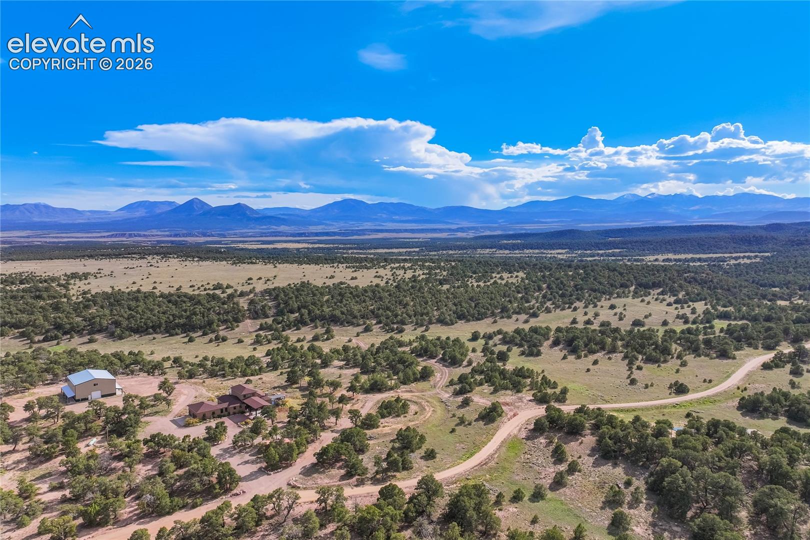 Expansive natural landscape featuring scattered trees, a single-story residence, and a separate outbuilding, all set against a distant mountain range