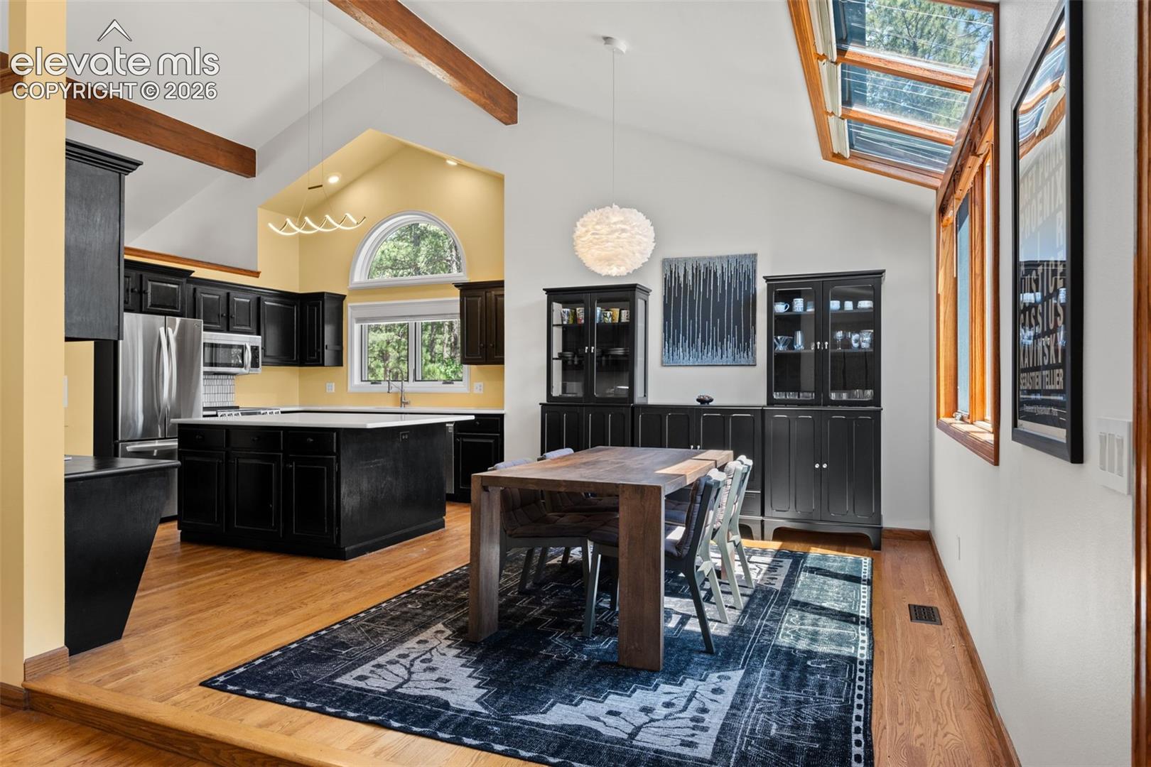 Vaulted ceilings featuring exposed wood beams, wood-finish flooring, black cabinetry, a central island with a white countertop, and multiple skylights