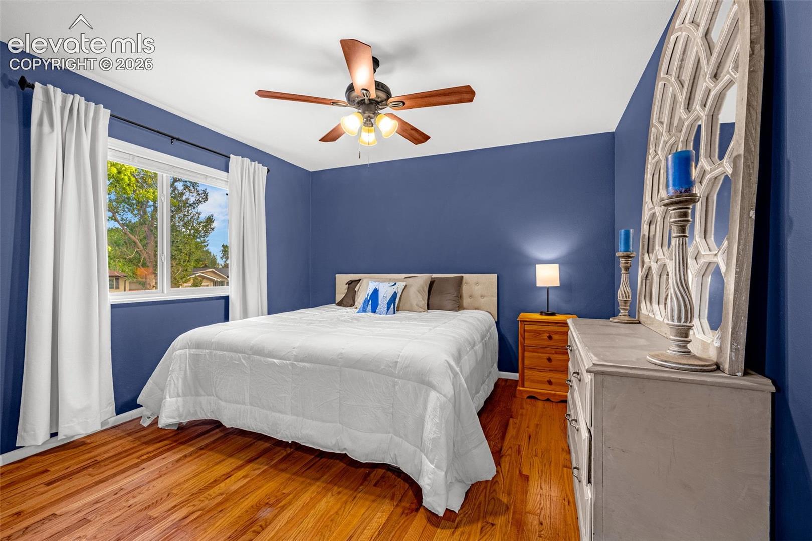 Primary bedroom with king size bed, hardwood flooring, ceiling fan with integrated lighting, and a large window