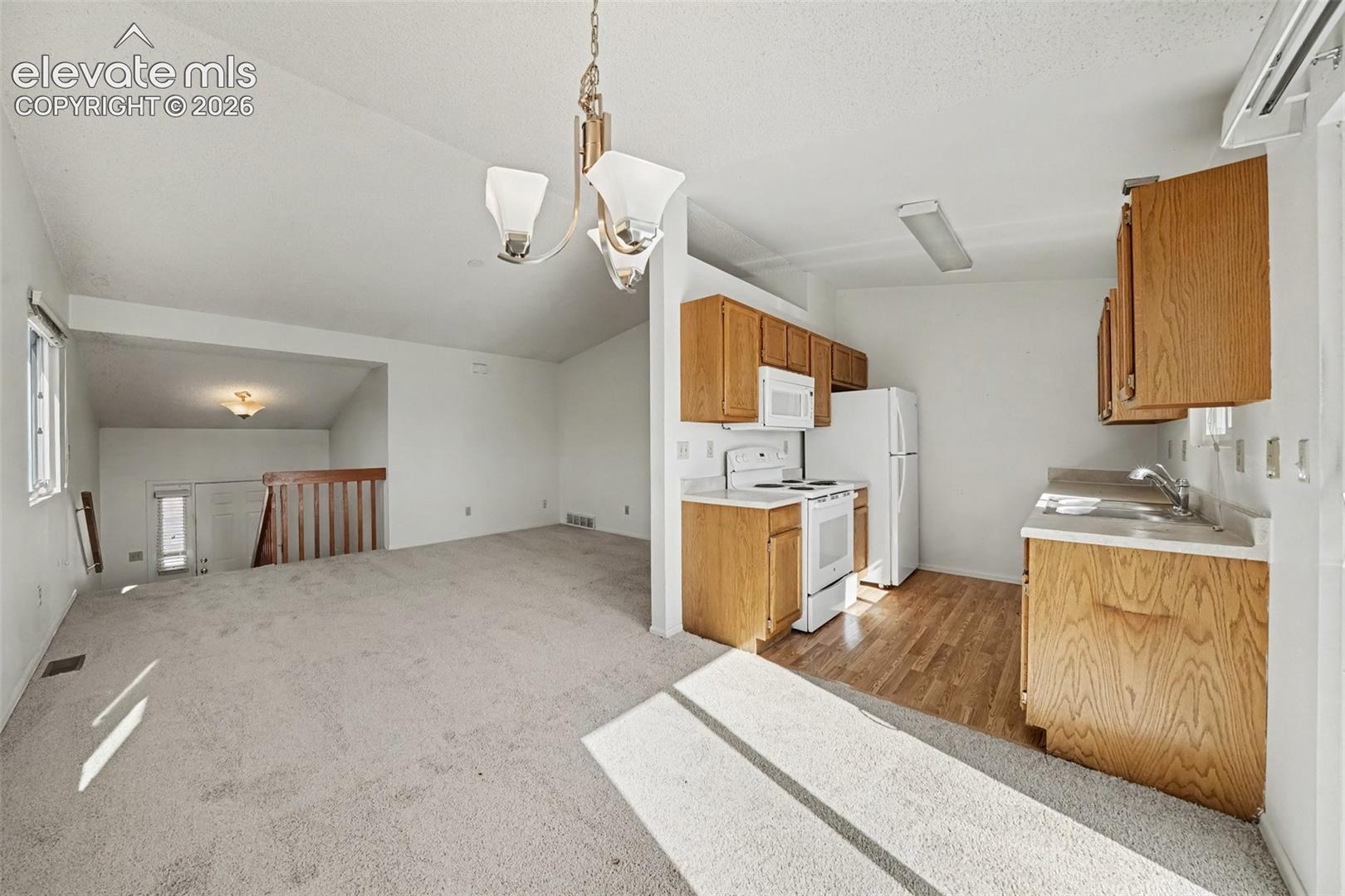 Kitchen featuring wood cabinetry, white appliances, and wood-finish flooring, set against a carpeted area with a vaulted ceiling, multi-arm chandelier, and wood railing