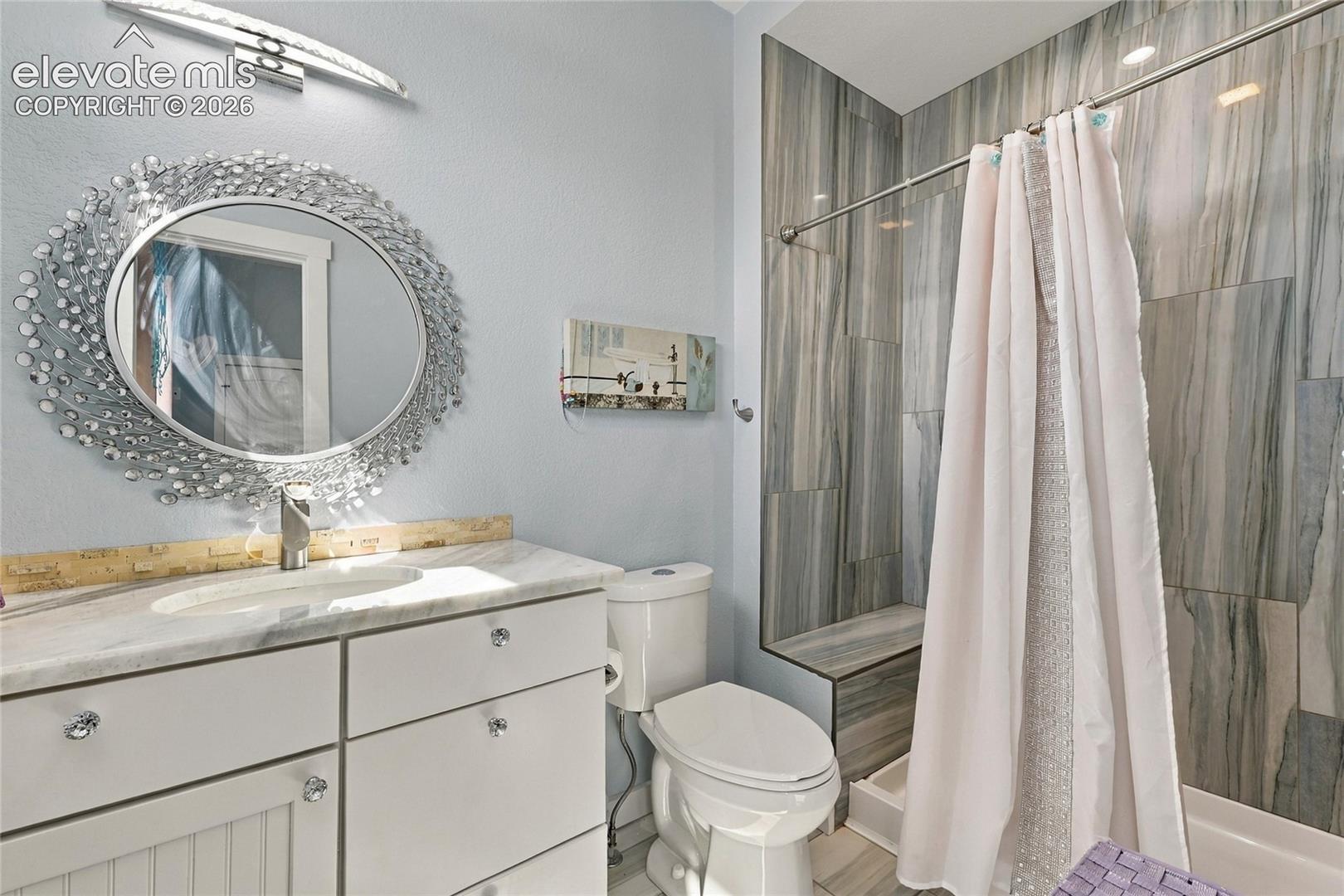 Bathroom vanity featuring white cabinetry, stone-finish countertop, oval basin, and a decorative circular mirror, with a shower surround clad in large-format patterned tile