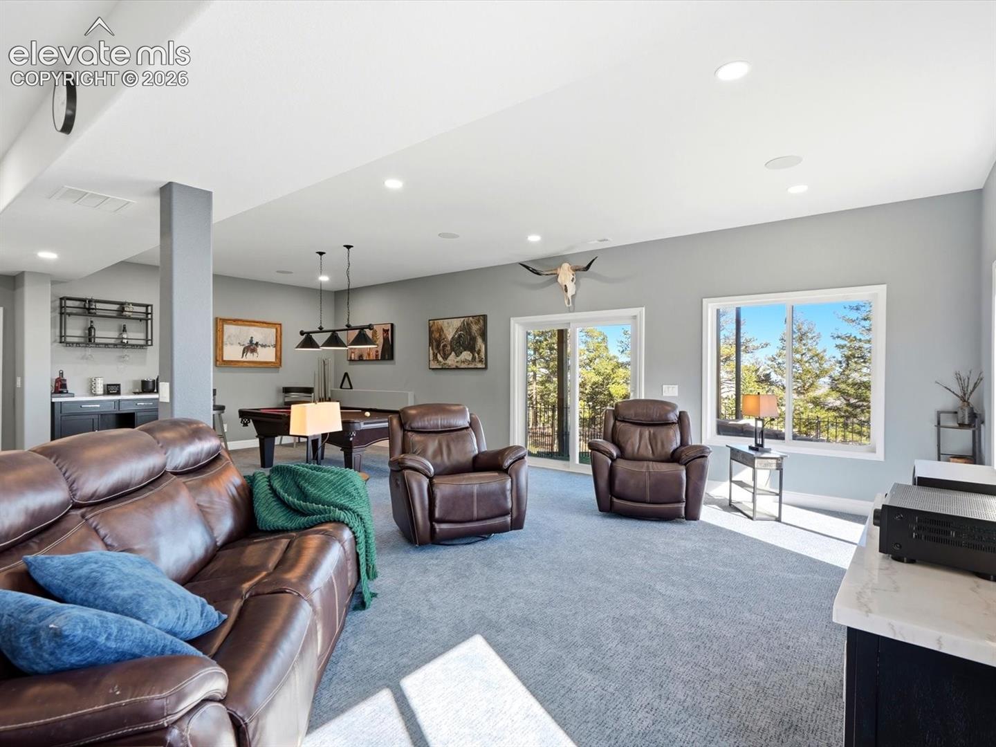 Open-concept interior featuring recessed lighting, a built-in wet bar with dark cabinetry, sliding glass doors, and large windows with exterior forest scenery