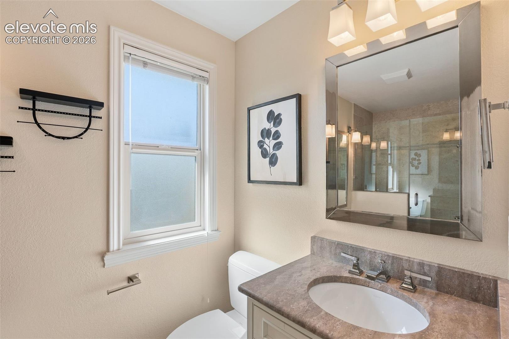 Stone countertop with an integrated undermount sink, multi-light vanity fixture, large rectangular mirror, and frosted glass window