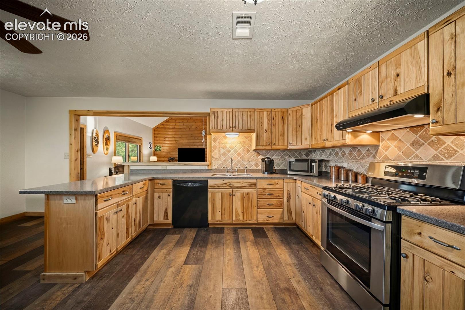 Kitchen featuring custom natural wood cabinetry, multi-tonal wood-finish flooring, stone-tiled backsplash, and stainless steel appliances