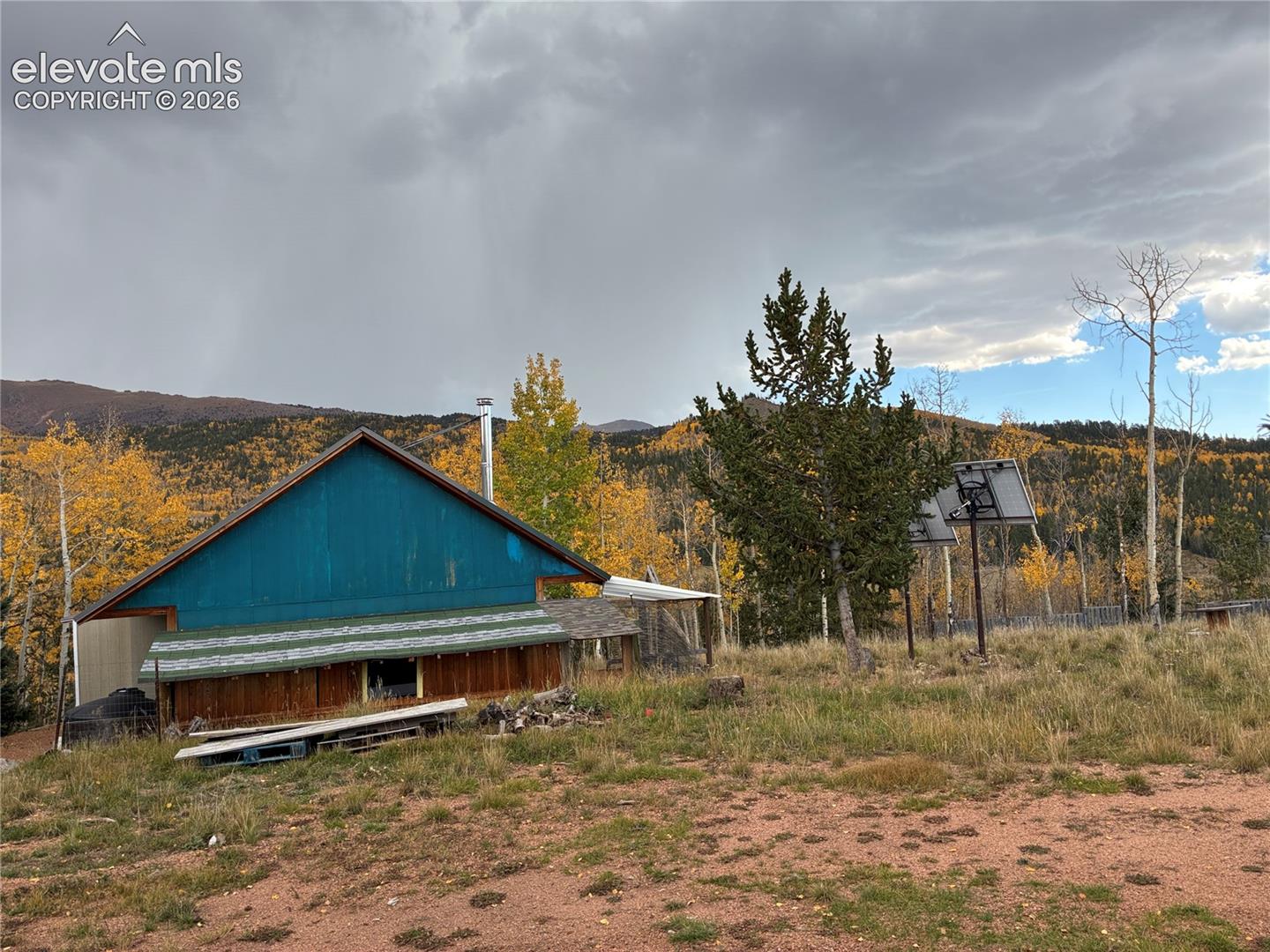 Gable-roof exterior with vertical siding and metal chimney, set against a mountain backdrop with native aspen trees and installed solar panels