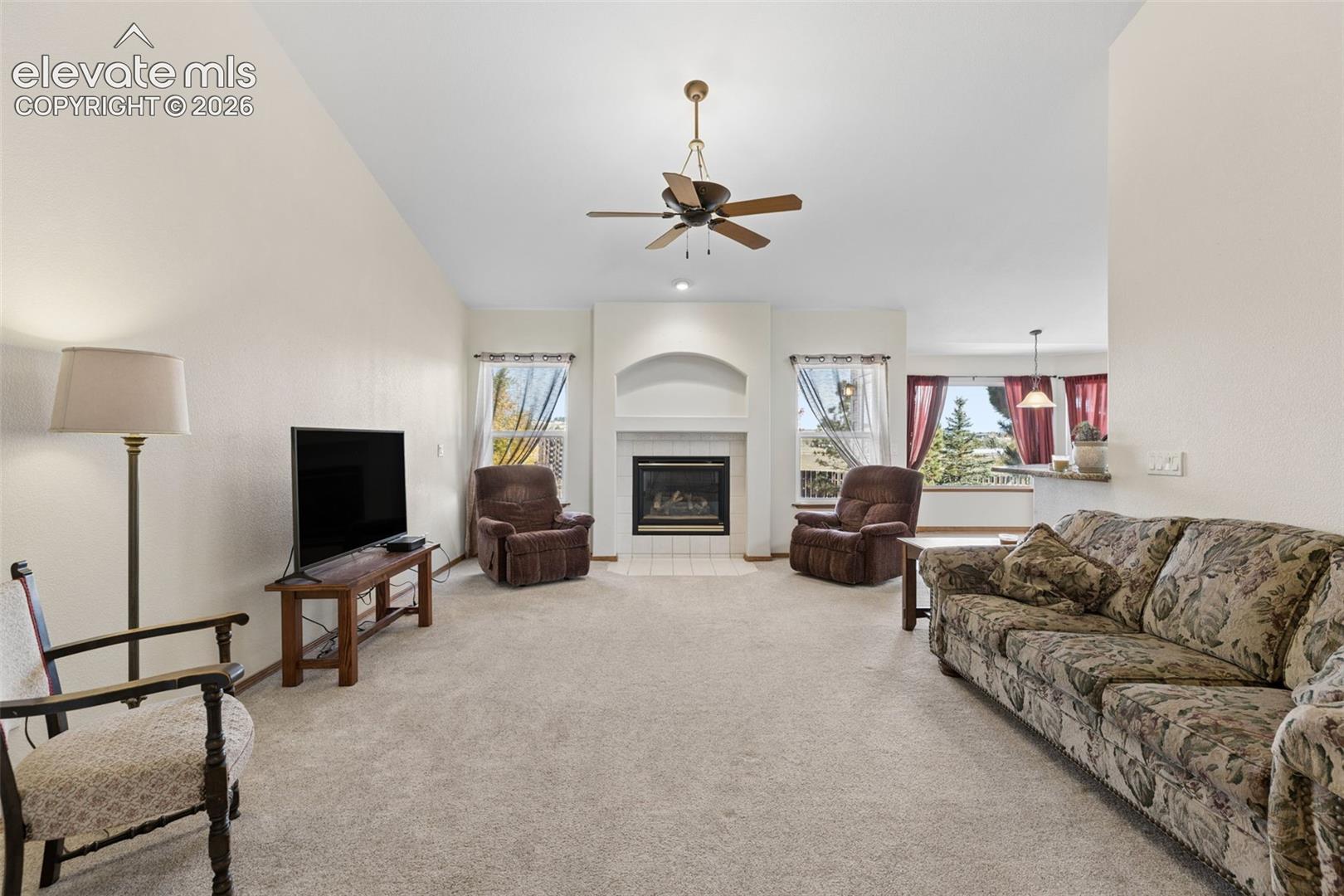 Living area featuring vaulted ceilings, a central fireplace with a tile hearth and arched alcove, neutral wall-to-wall carpeting, and multiple windows