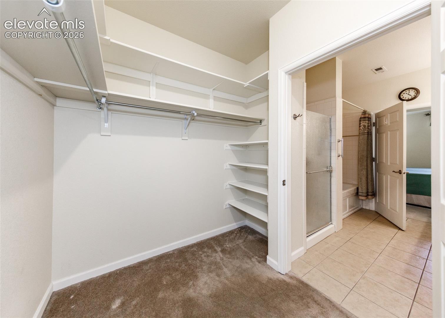 Walk-in closet featuring white tiered shelving and integrated hanging rods, offset by a bathroom with tiled flooring, a glass-enclosed shower, and a soaking tub