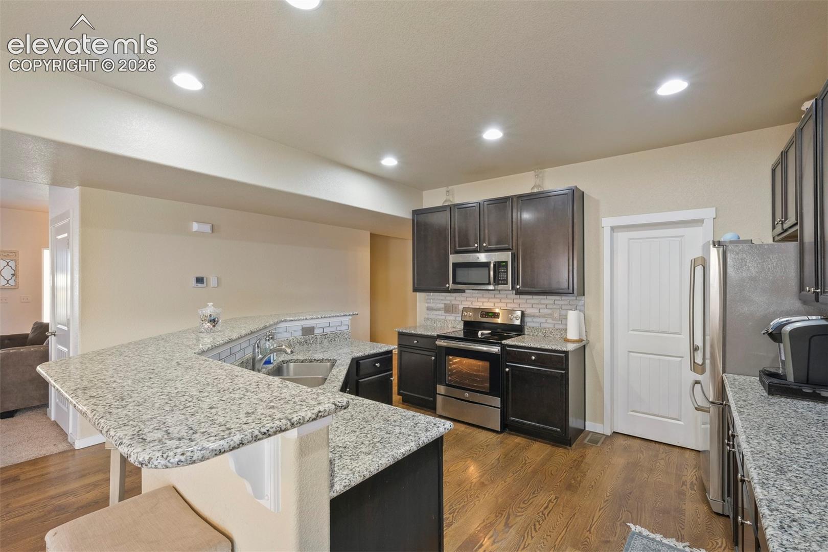 Modern kitchen featuring granite countertops, wood-finish flooring, dark wood cabinetry, recessed lighting, and stainless steel appliances