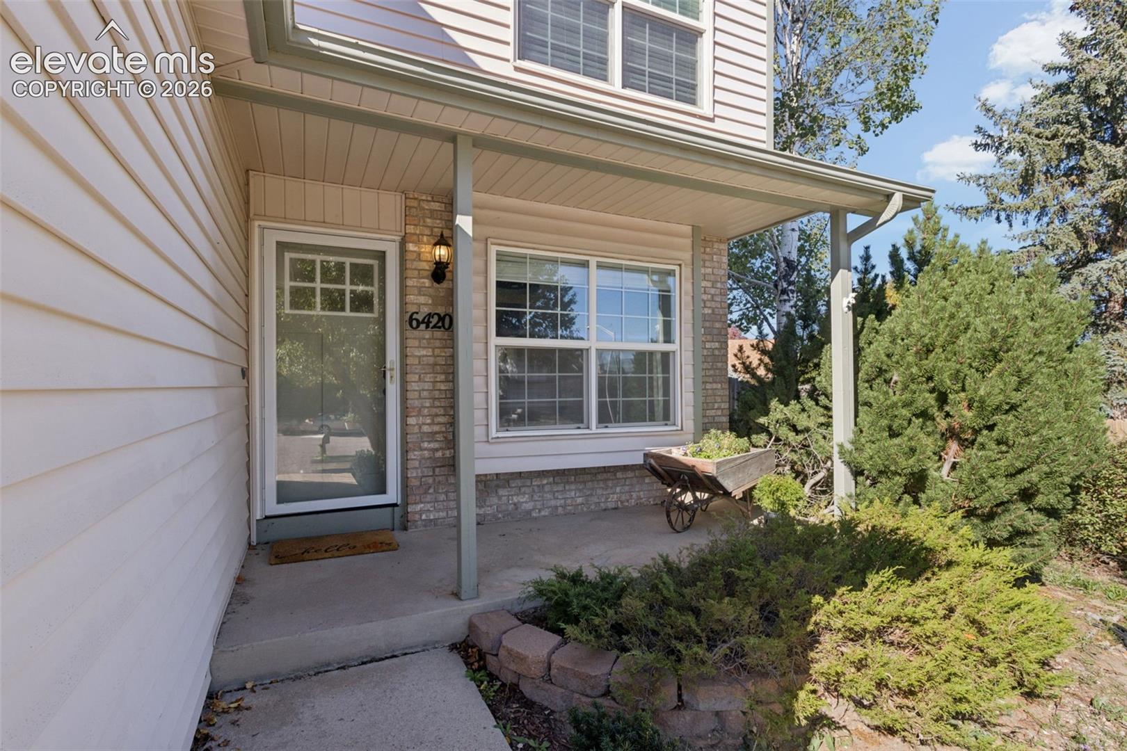 Covered front porch with concrete walkway, brick accents, and mature evergreen landscaping