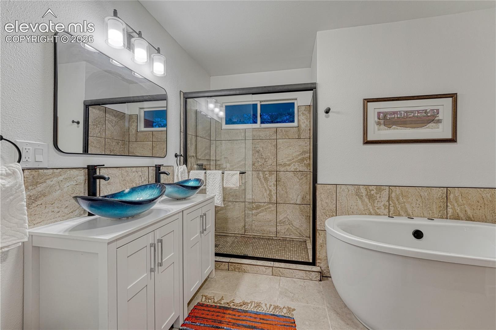 Double vanity with blue glass vessel sinks, walk-in shower with tiled surround, and freestanding soaking tub