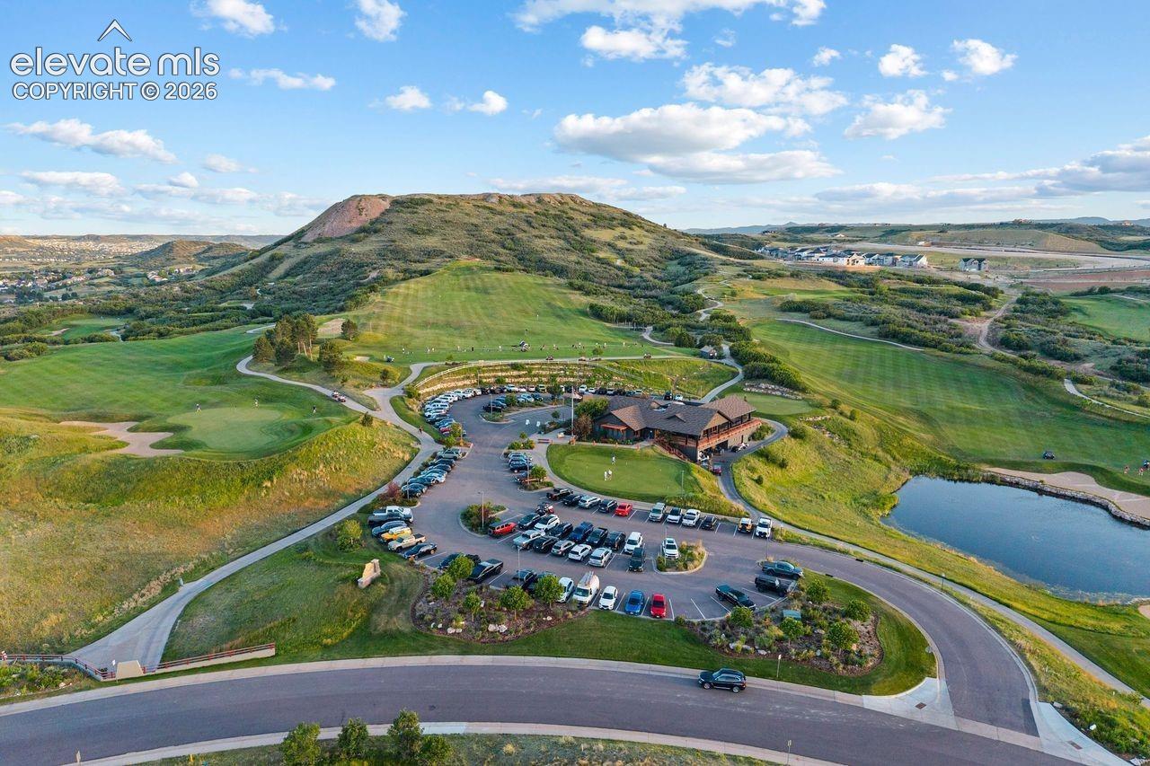 Aerial perspective of a clubhouse featuring extensive hardscaped parking, a large water feature, and rolling terrain set against a prominent mountain backdrop