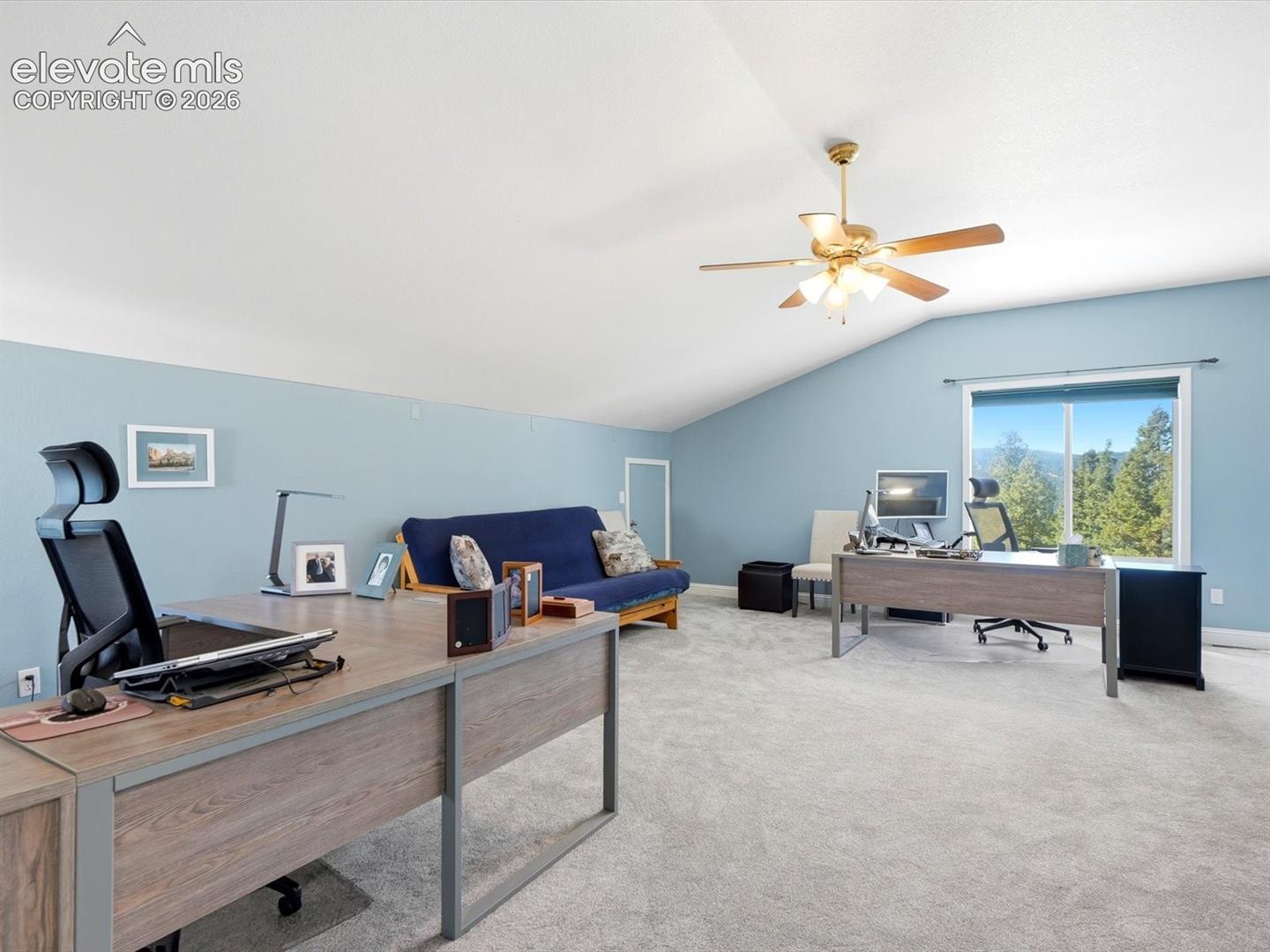 Vaulted ceiling featuring a central multi-blade light fixture, neutral wall-to-wall carpeting, and a large window with a forest backdrop