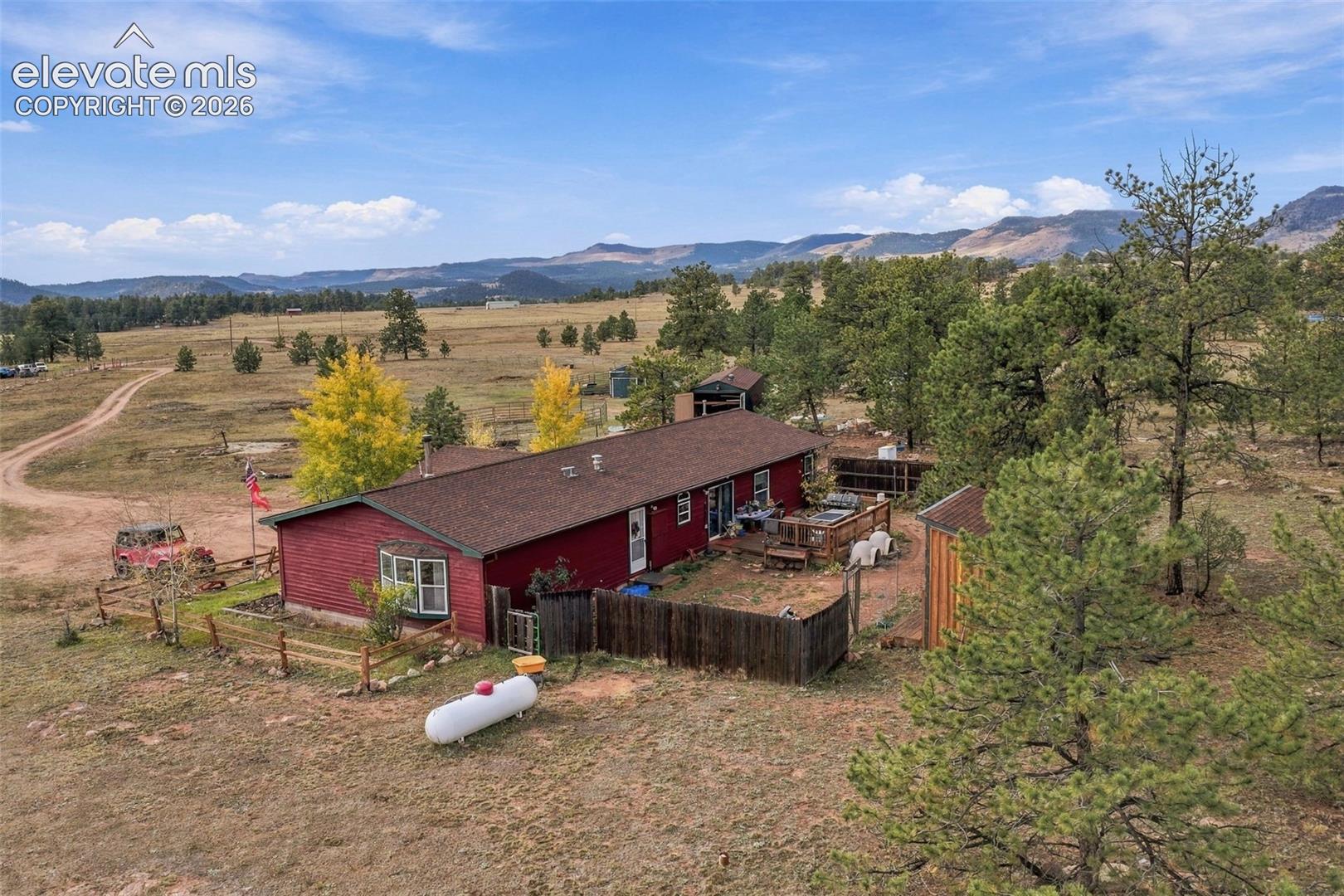 Single-story residence featuring horizontal siding, a bay window, shingle roofing, wooden perimeter fencing, and an attached deck, set against a mountainous backdrop