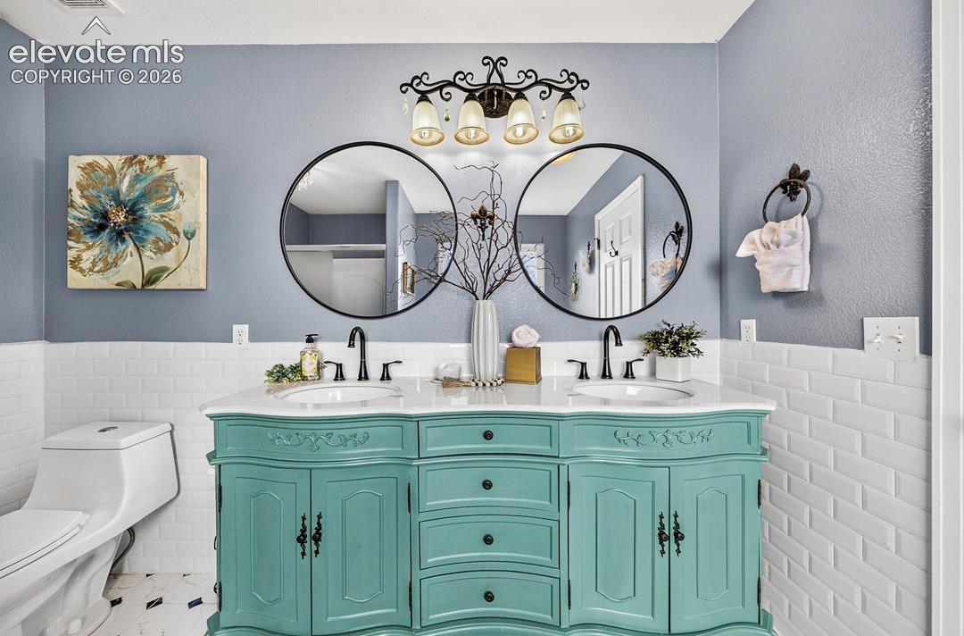 Dual-sink vanity featuring a painted cabinet finish, white countertop, and dark metal hardware, complemented by twin round mirrors, subway tile wainscoting, and decorative four-light vanity fixture