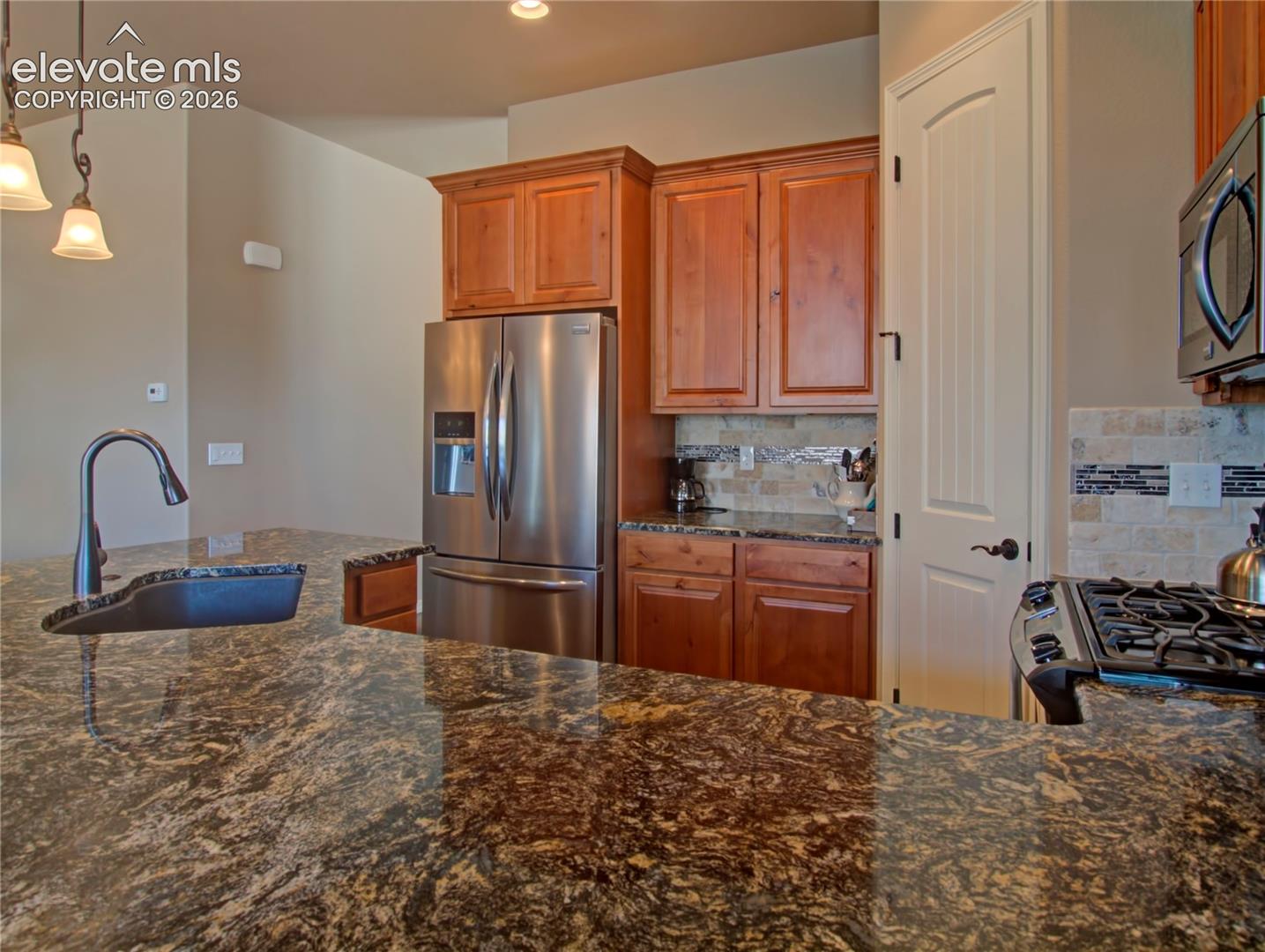 Spacious kitchen featuring expansive stone countertops, wood cabinetry, stainless steel appliances, a tiled backsplash, granite counters and pendant lighting