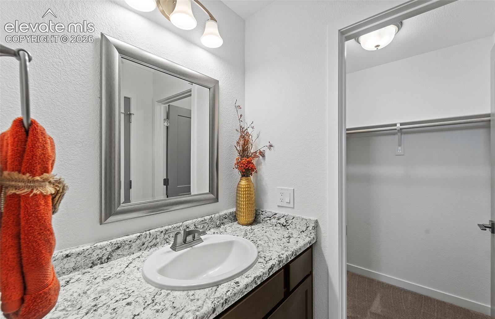 Bathroom vanity featuring a stone-pattern countertop, undermount sink, metallic faucet, framed wall mirror, and three-light vanity fixture, including a built-in closet with shelving and hanging rod