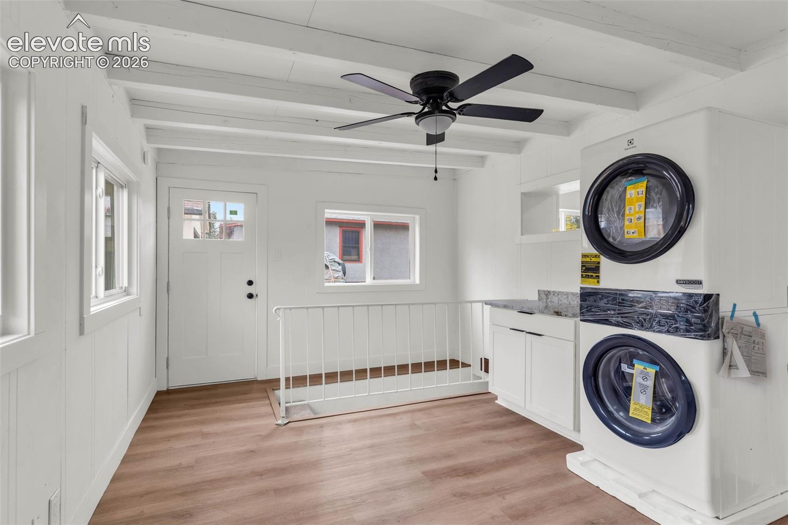 Wood-finish flooring, white exposed ceiling beams, built-in cabinetry with stone-finish countertops, stacked laundry unit, and a ceiling fan