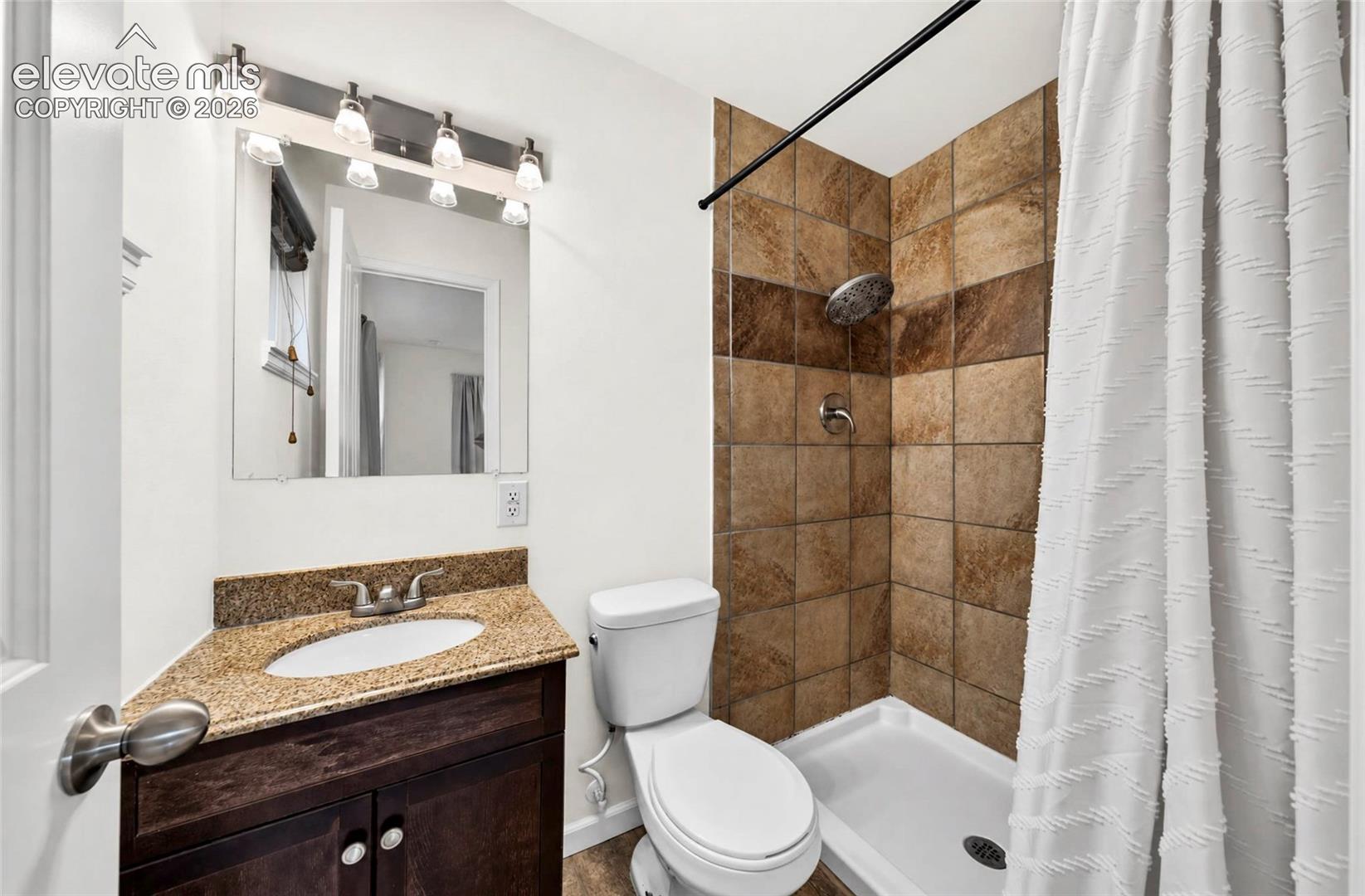 Dark wood-finish vanity with a stone-composite countertop, integrated sink, tile-surround shower, and multi-bulb wall-mounted vanity lighting