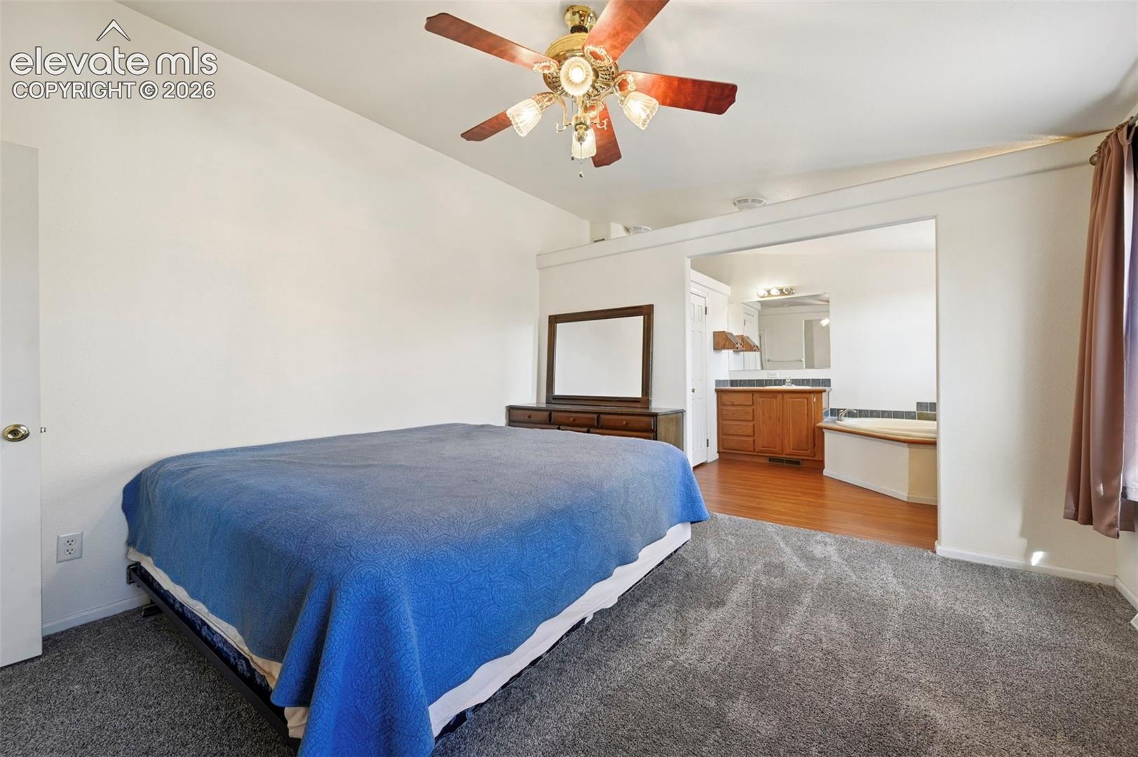 Room featuring carpeted flooring, vaulted ceilings, and a ceiling fan with integrated lighting, with a wood-finish vanity and platform tub visible in the secondary area