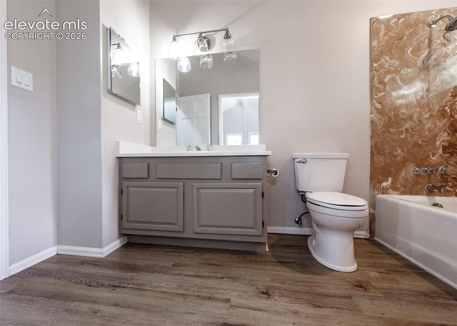Wood-finish flooring, single vanity with shaker-style cabinetry, white countertop, and a built-in bathtub with a patterned surround