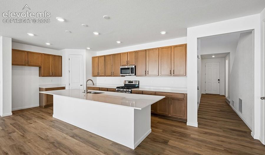 Kitchen featuring wood-finish cabinetry, white stone-composite countertops, a central island with a stainless steel undermount sink, recessed lighting, and wood-finish flooring