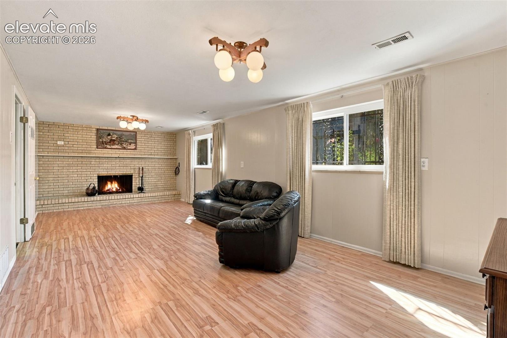 Living area featuring wood-finish flooring, a full-wall brick fireplace with an integrated hearth, and dual multi-light ceiling fixtures
