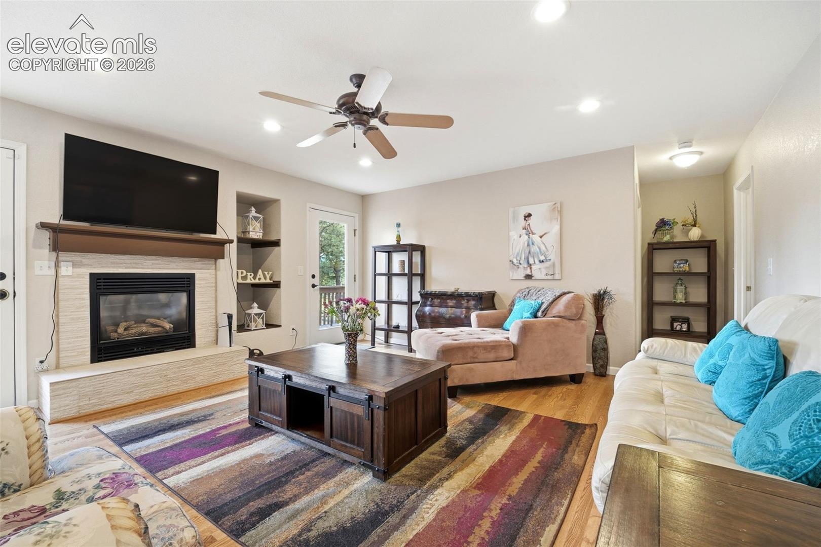 Living room featuring wood-finish flooring, a fireplace with a light-toned surround and wood mantel, recessed lighting, and a ceiling fan