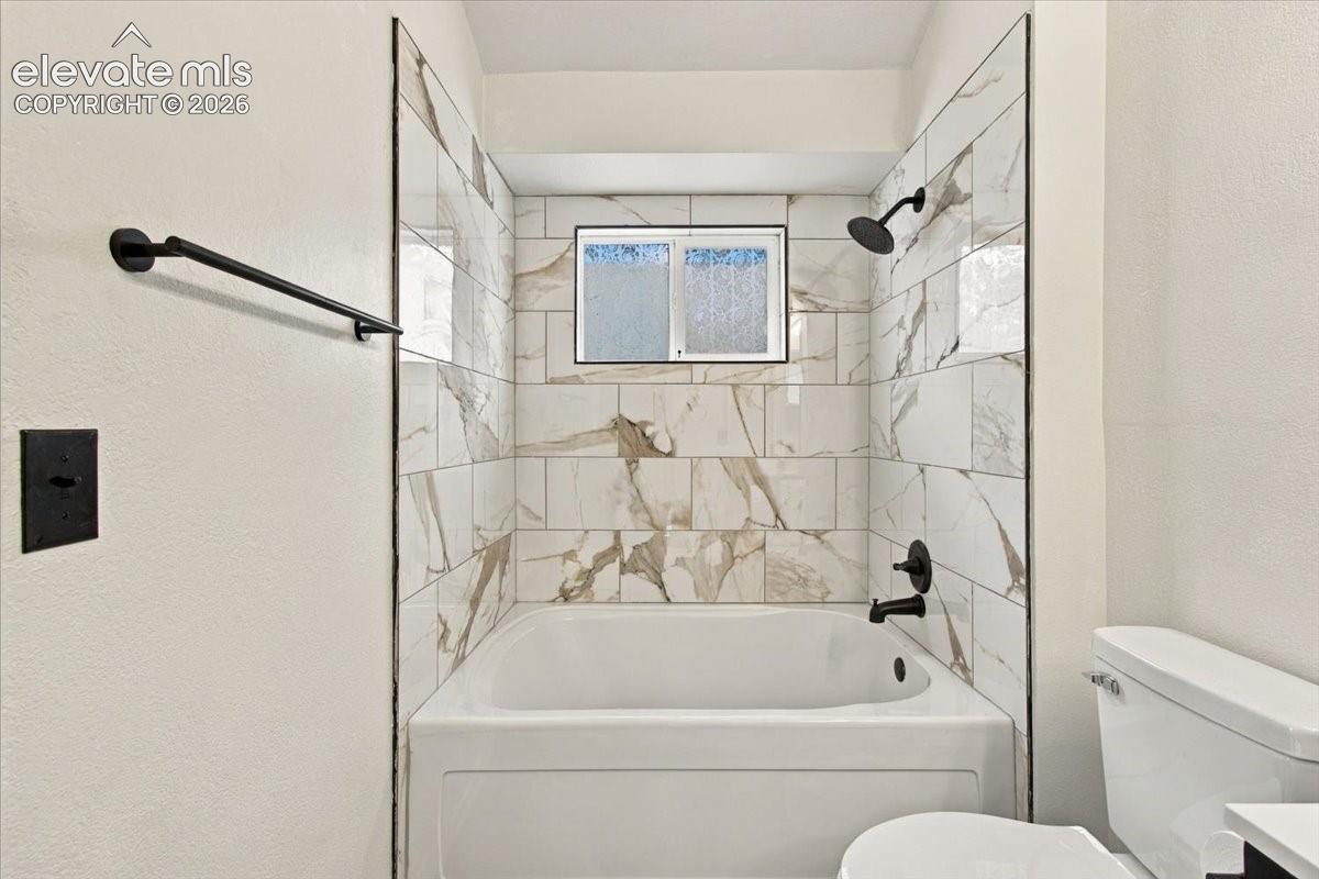 Bathtub enclosure featuring large-format stone-pattern tile surround, matte black rainfall showerhead, matching matte black faucet, and built-in matte black towel bar