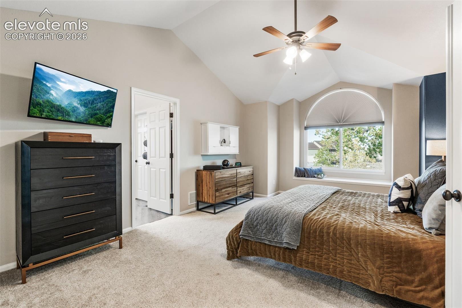 Vaulted ceilings with an integrated ceiling fan, carpeted flooring, and a large arched window with a built-in window seat