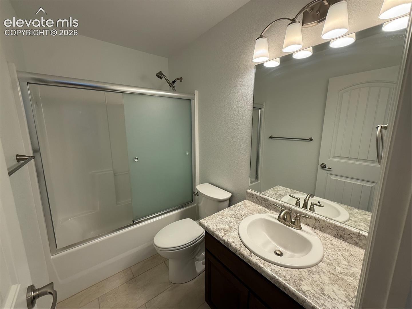 Bathroom featuring a vanity with a stone-patterned countertop, an integrated oval sink, a shower-tub combination with sliding frosted glass doors, and a full-width wall mirror with multi-light fixture
