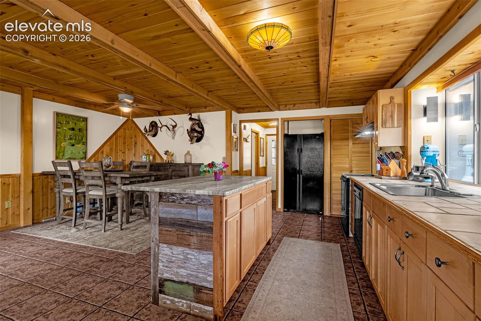Kitchen featuring exposed wood beam ceilings, a central island with natural stone-finish countertop, tile flooring, and light wood cabinetry