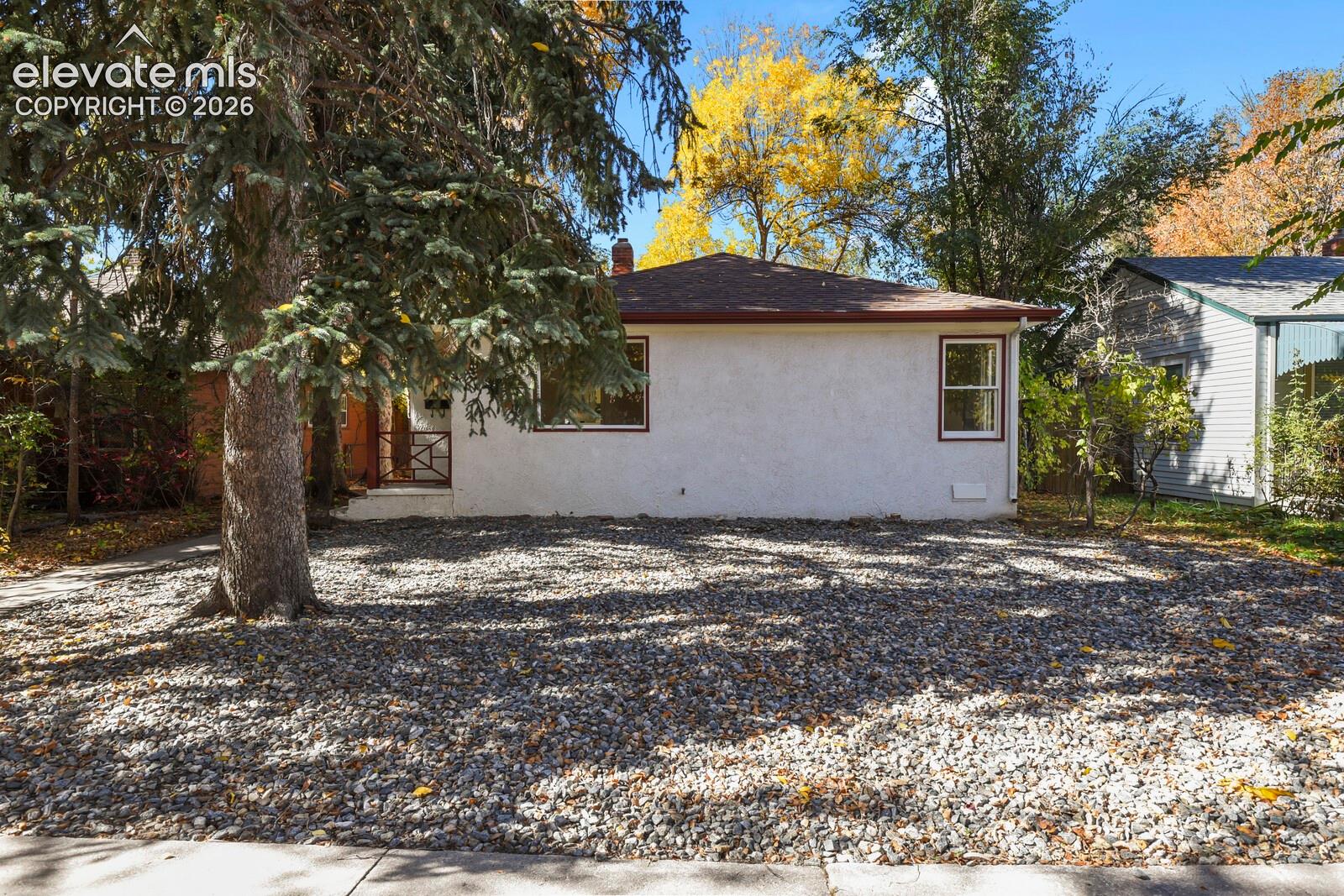 Single-story residence featuring a light-colored stucco exterior, decorative metal porch railing, shingled roof, and gravel front landscaping with a mature evergreen tree