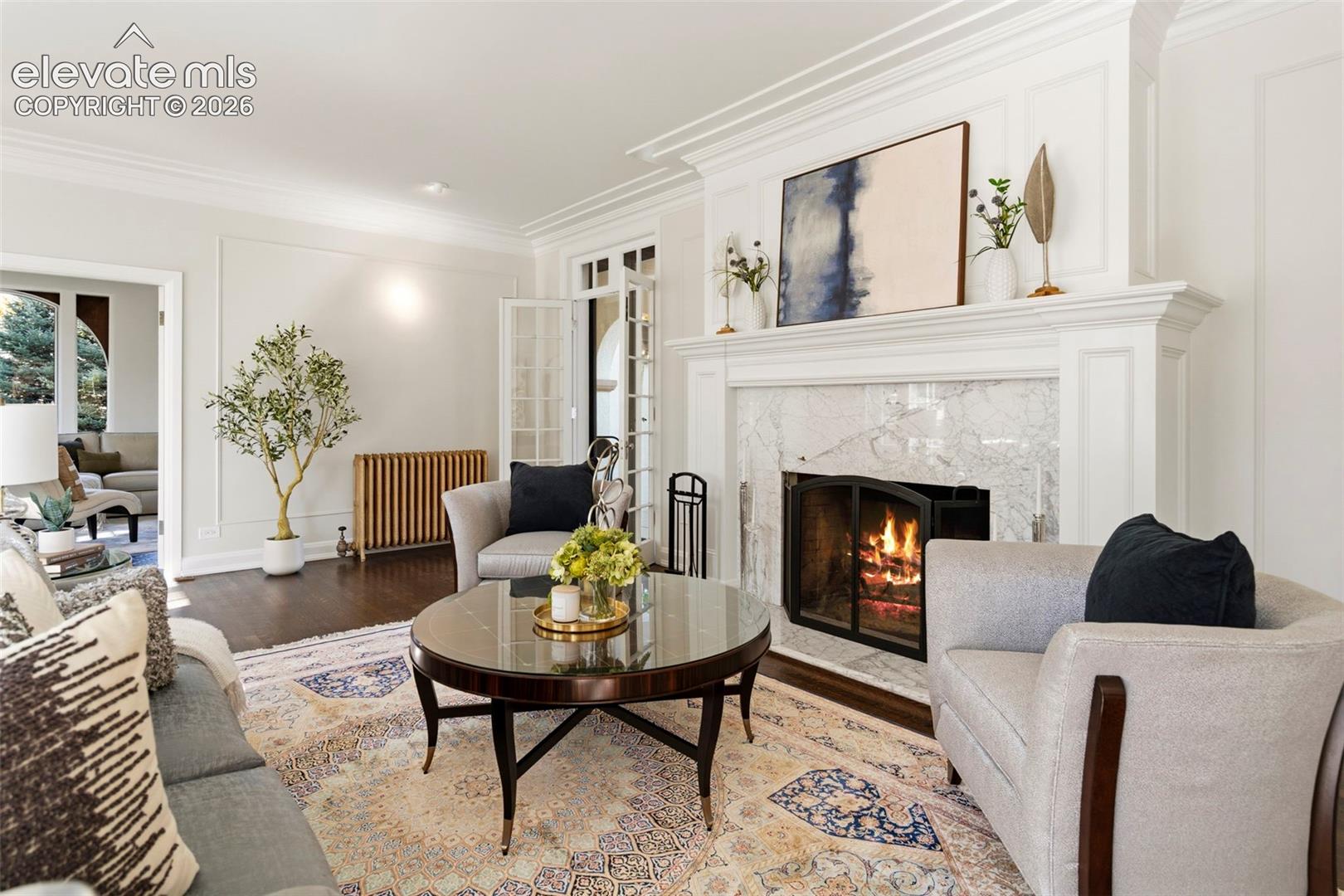 Living area featuring a fireplace with stone surround, custom millwork mantel, crown molding, paneled walls, and dark wood-finish flooring