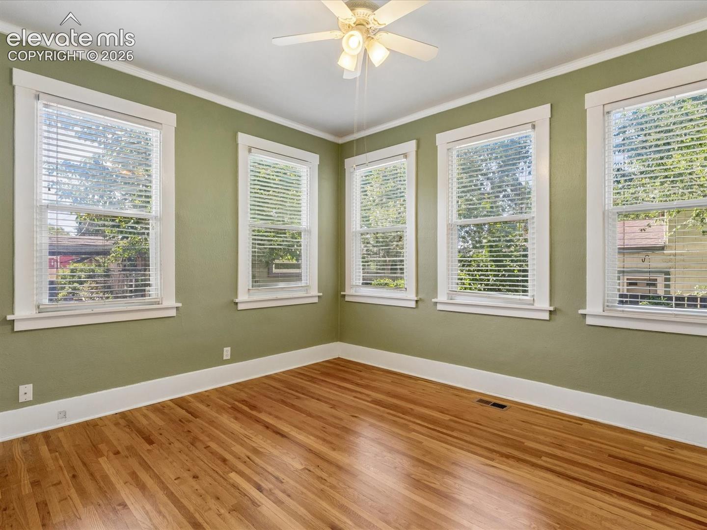 Spacious room featuring hardwood flooring, multiple windows with white blinds, crown molding, and a ceiling fan with integrated lighting
