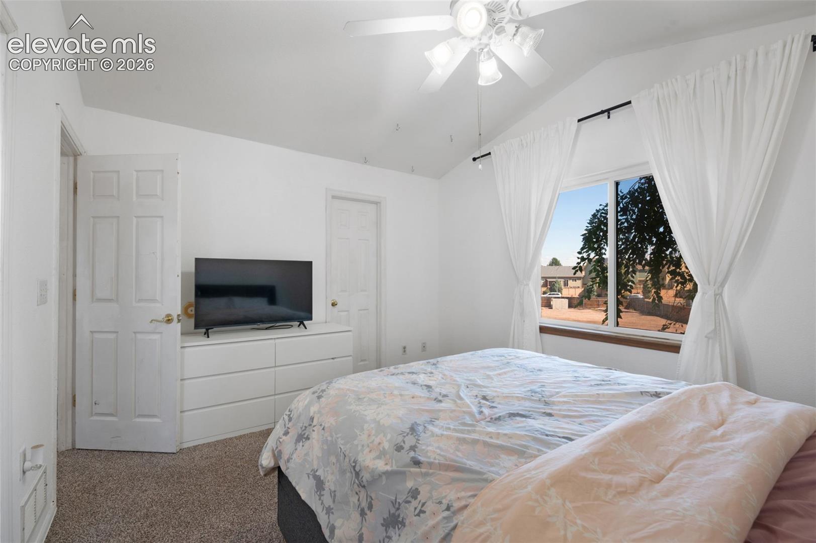 Carpeted bedroom featuring a vaulted ceiling with a ceiling fan, a large window with white curtains, and a six-panel interior door