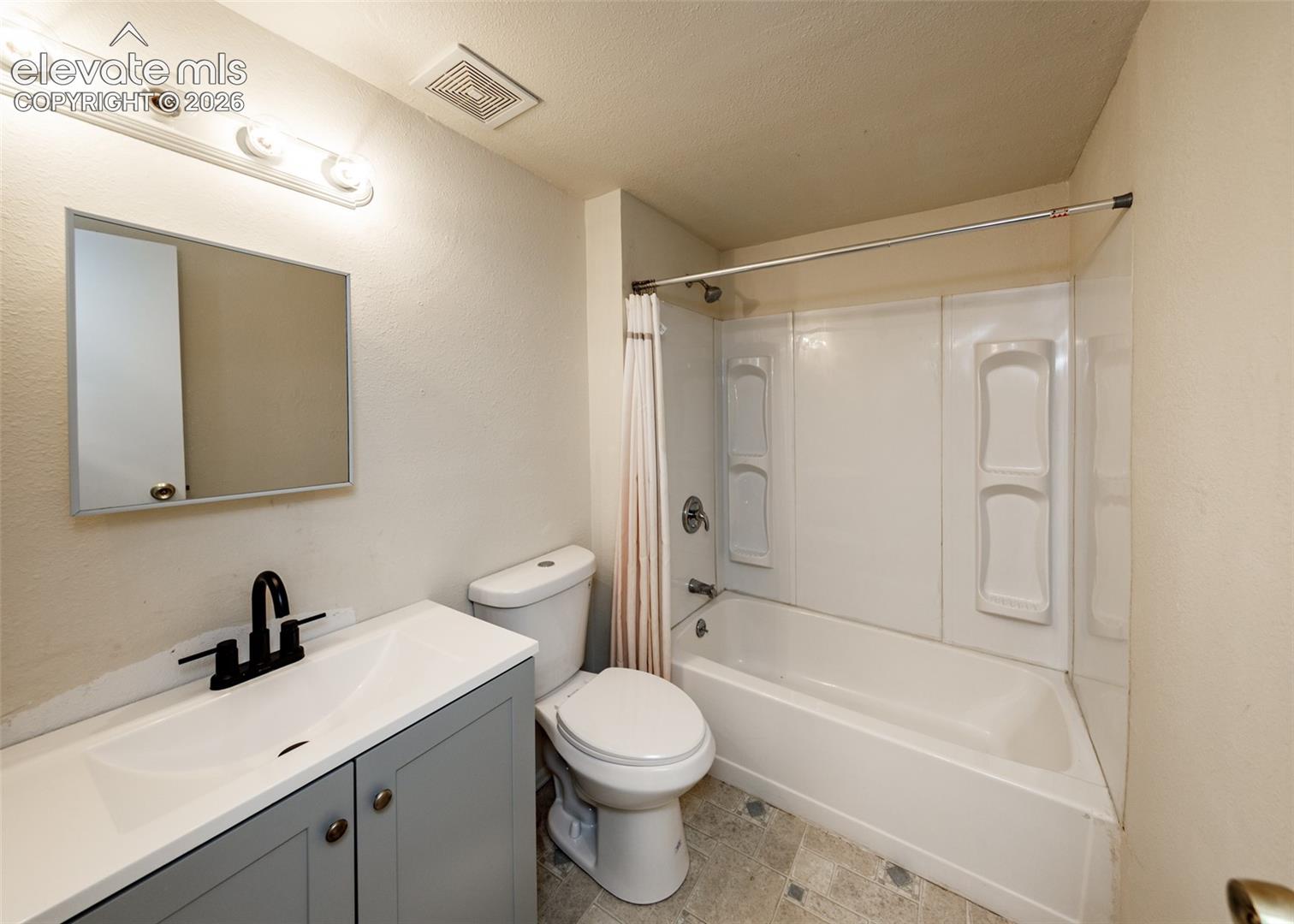 Bathroom featuring a grey vanity cabinet with a stone-top integrated sink, matte black faucet, wall-mounted mirror, and a tub-shower combination with built-in shelving