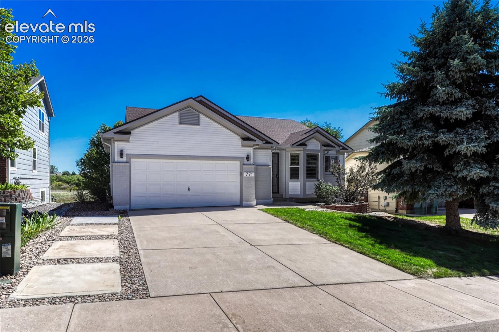 Gray single-story home with a two-car garage, broad driveway, and large evergreen tree in the front yard.