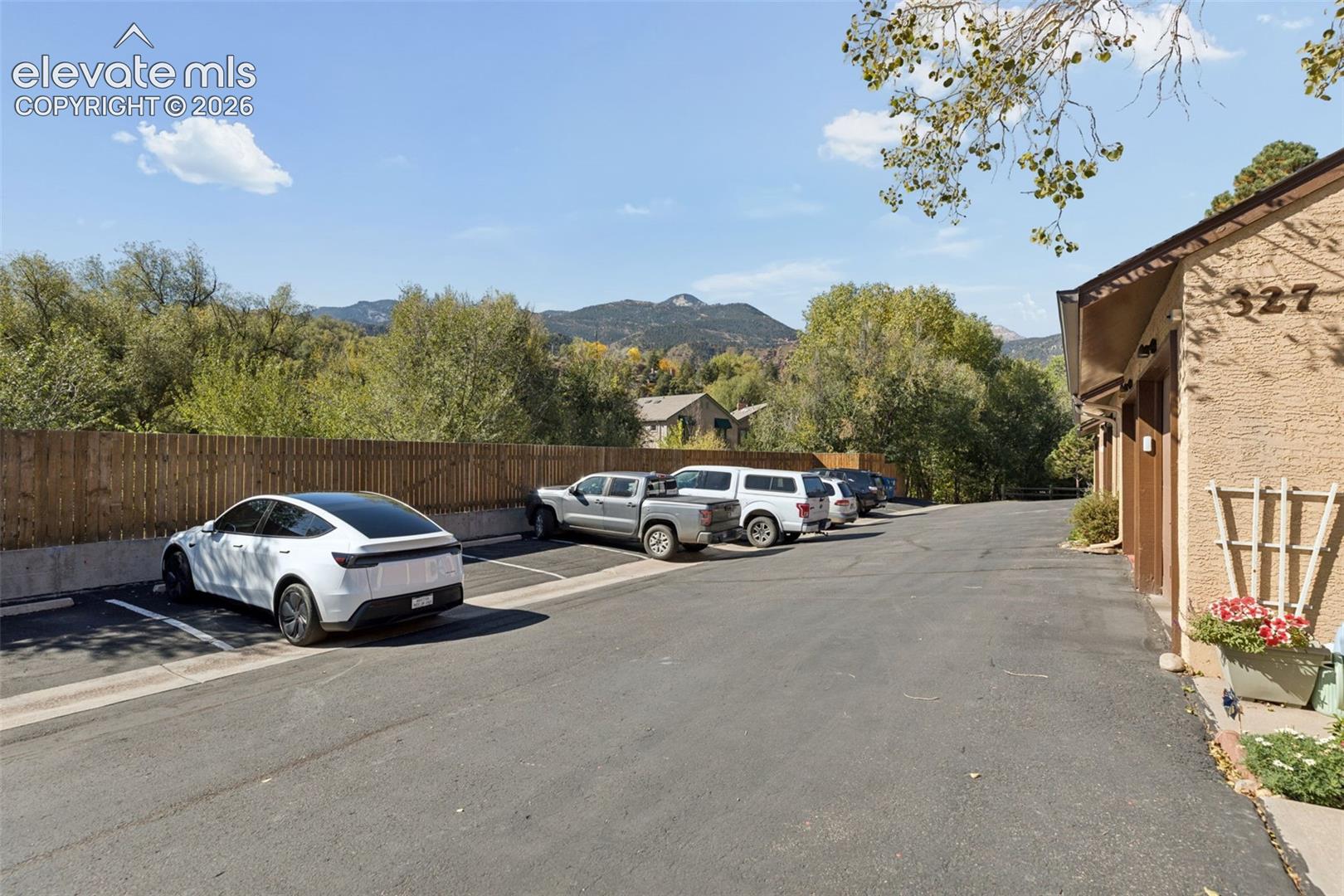 Paved parking area, stucco exterior, privacy fencing, mountain backdrop, and mature trees