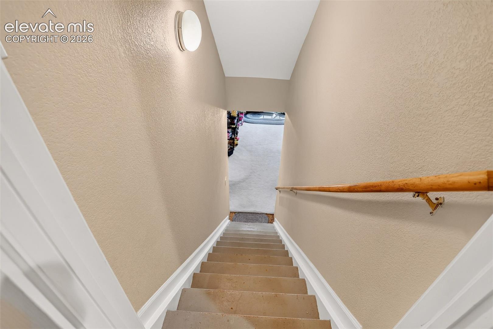 Staircase going to basement with light beige textured walls, white trim, wood handrail, and overhead flush-mount lighting