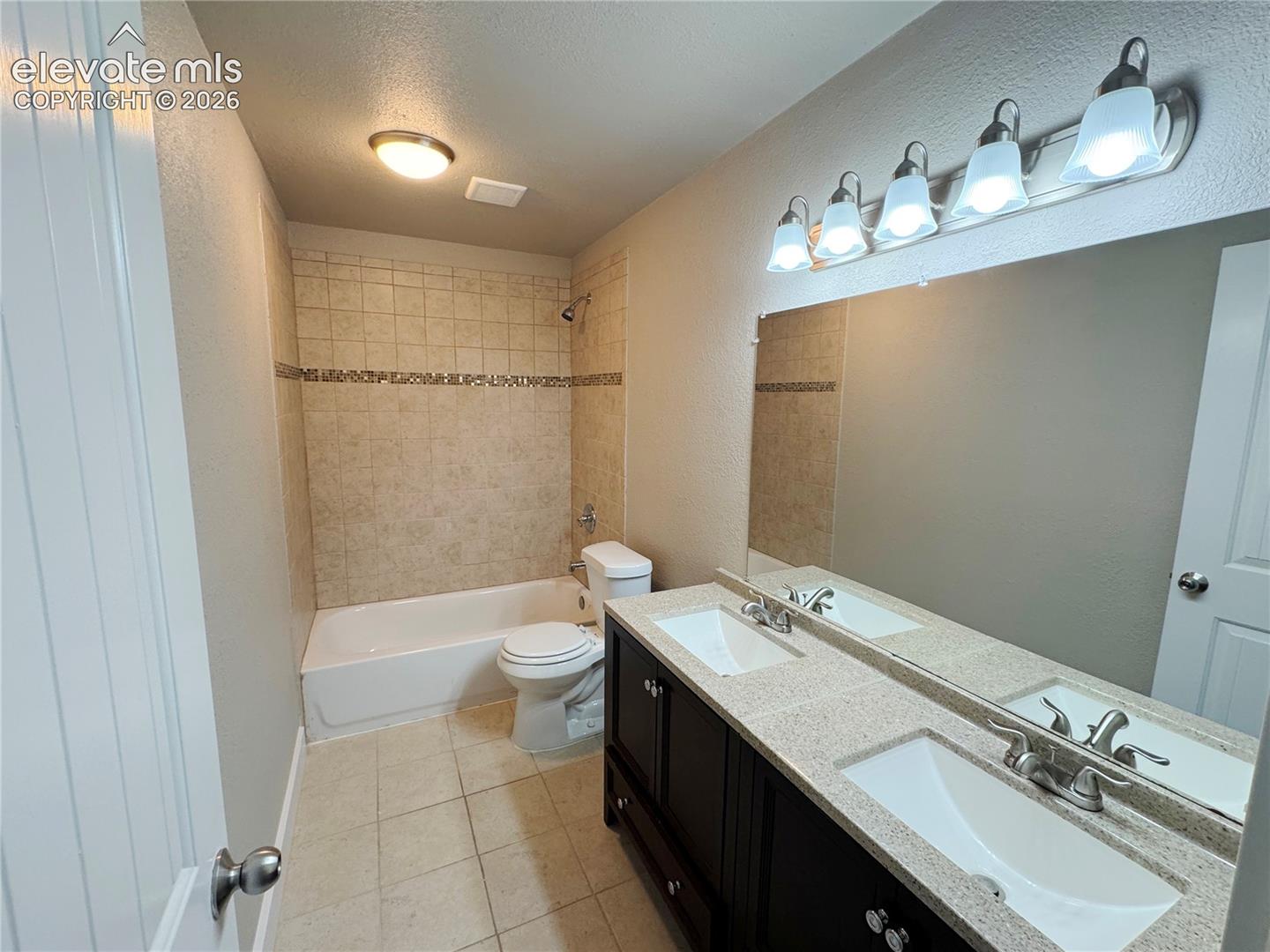 Double vanity featuring a light-toned solid surface countertop with dual integrated sinks, dark wood-finish cabinetry, a large wall-mounted mirror, five-light fixture, and a tub-shower combination with decorative tile surround