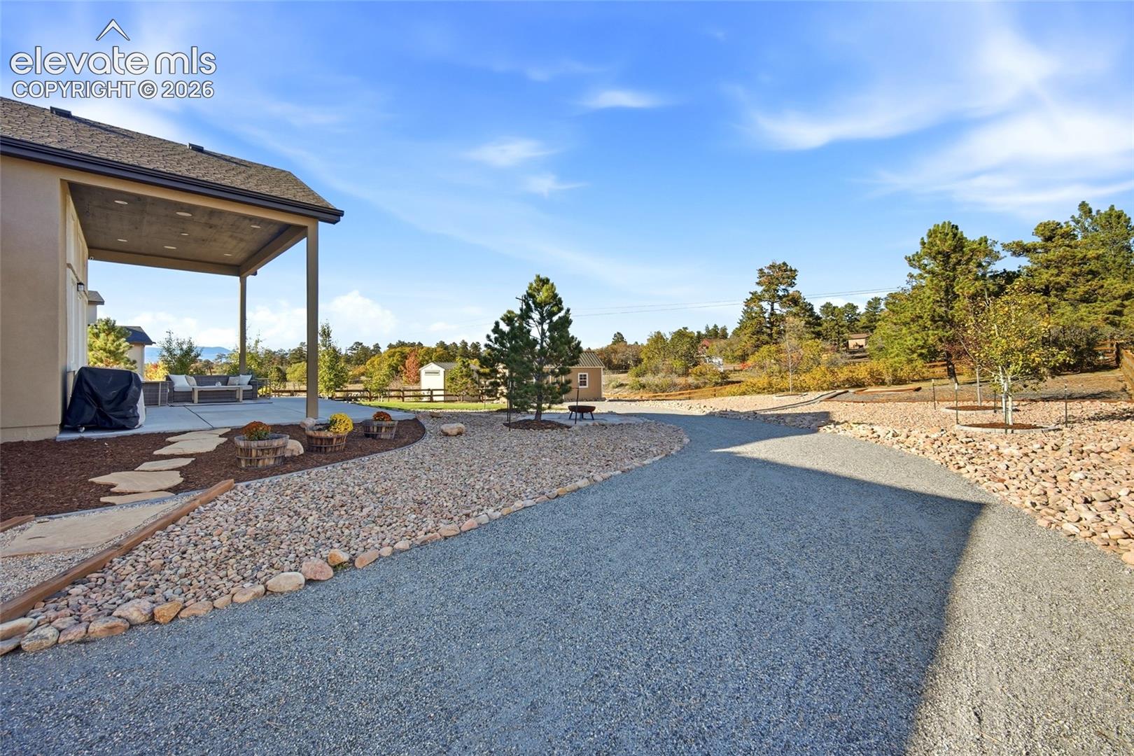 Covered concrete patio with recessed lighting, gravel driveway, stone-bordered mulch landscaping, and flagstone path