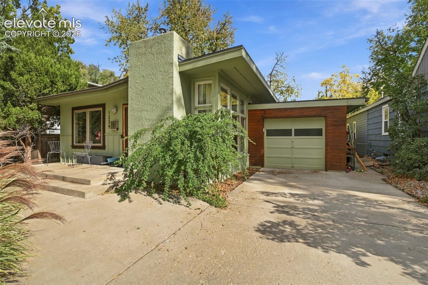 Mid-century architectural exterior featuring a prominent chimney, low-pitch roofline, integrated single-car garage with wood-accented siding, and a concrete front patio