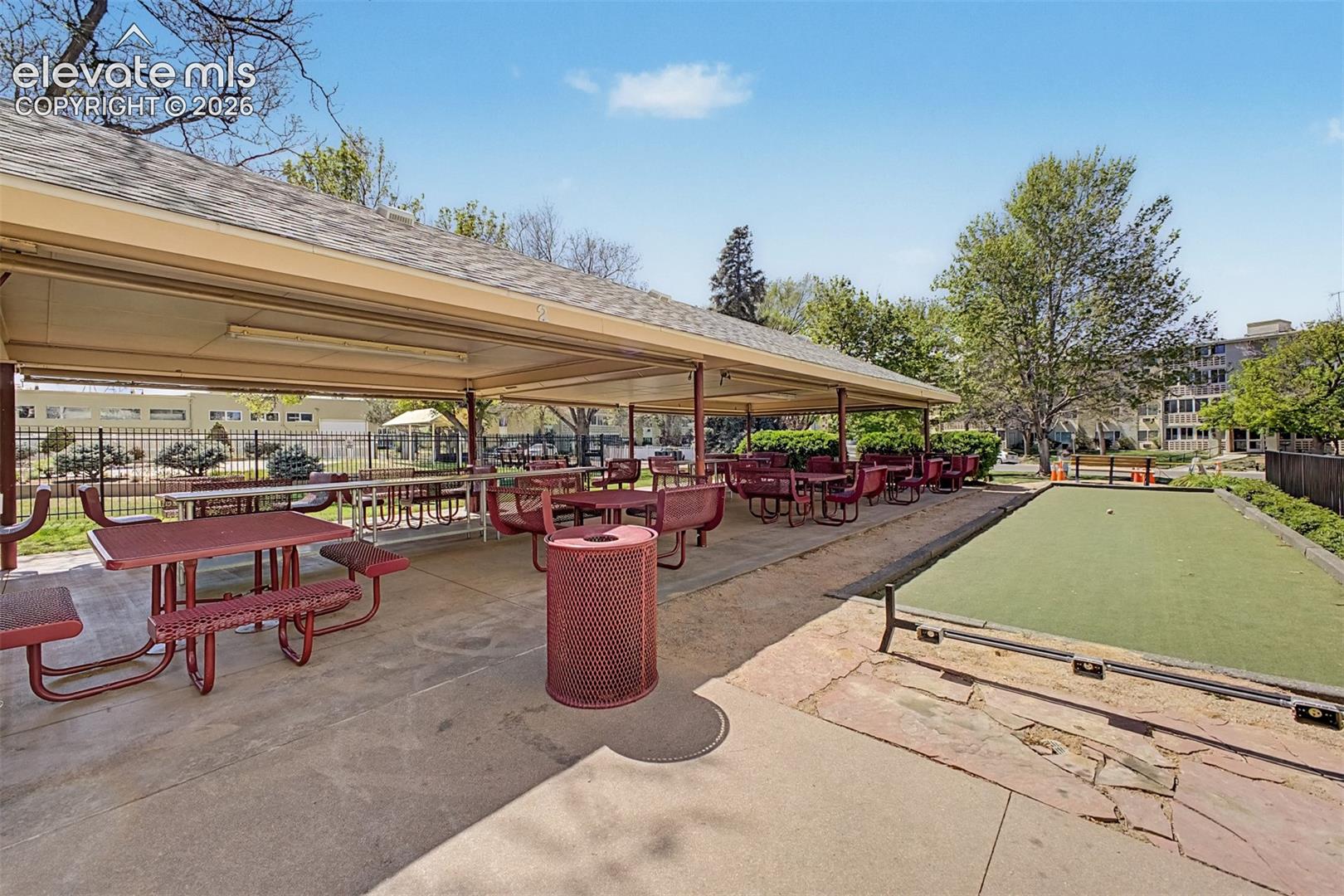 Covered outdoor patio area featuring multiple picnic tables with attached benches, a bocce ball court, and surrounding mature trees