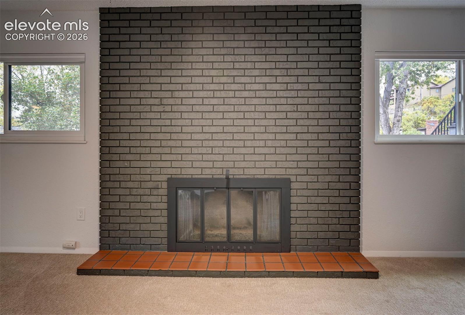 Floor-to-ceiling dark brick fireplace surround with a metal firebox insert and raised terracotta tile hearth, flanked by dual picture windows