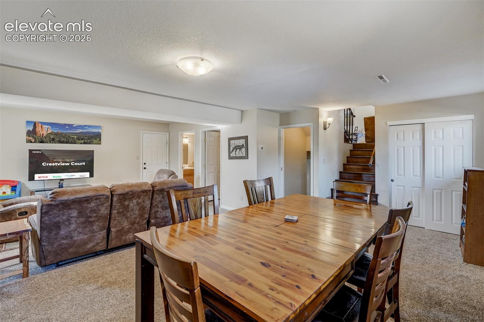 Carpeted living area featuring a wood-finish dining table, recessed lighting, and a wood staircase with a dark railing