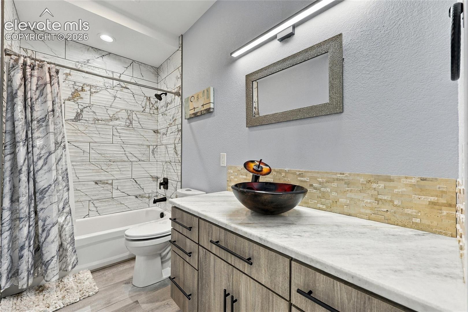 Bathroom vanity featuring a stone-finish countertop, wood-finish cabinetry, vessel sink, and stacked-stone backsplash, with floor-to-ceiling tiled shower surround