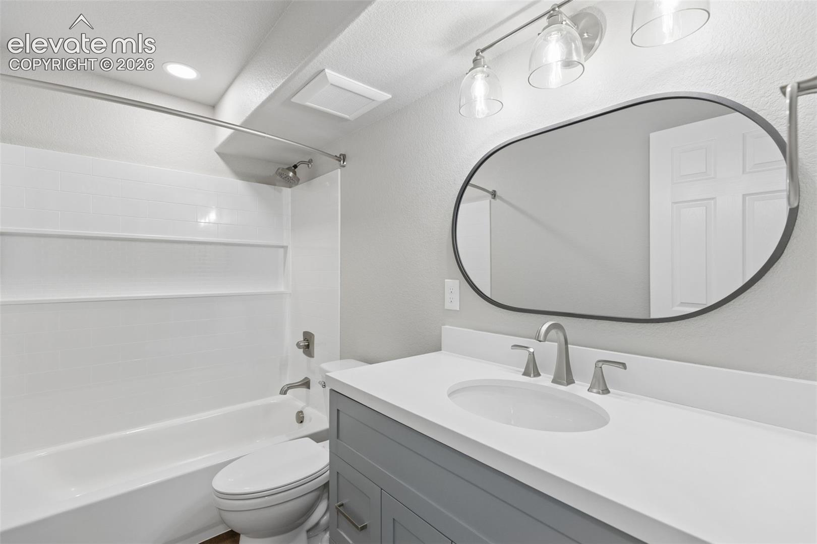 Bathroom featuring a grey vanity with a solid surface countertop, an oval-shaped mirror, a three-light fixture, and a tub-shower combination with white wall tile
